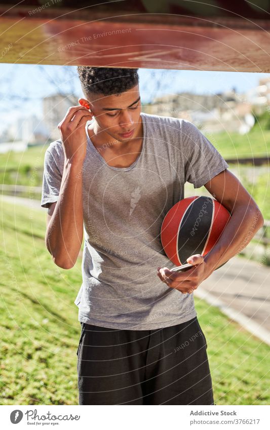 Streetball player with ball listening to music sportsman streetball smartphone earbuds using training break put on adjust athlete young workout basketball