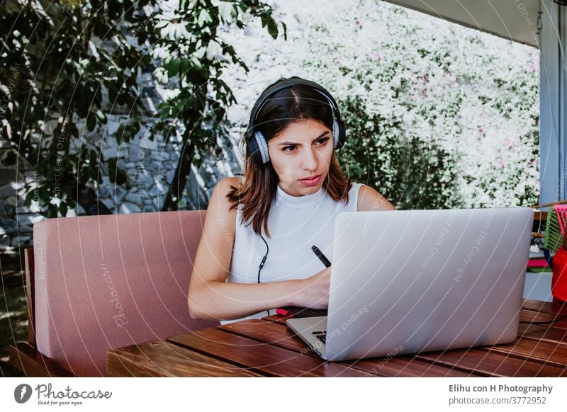 young latin woman sitting alone home office working on a laptop and listening to music on earphones, Mexican girl in Mexico working at home wireless browsing