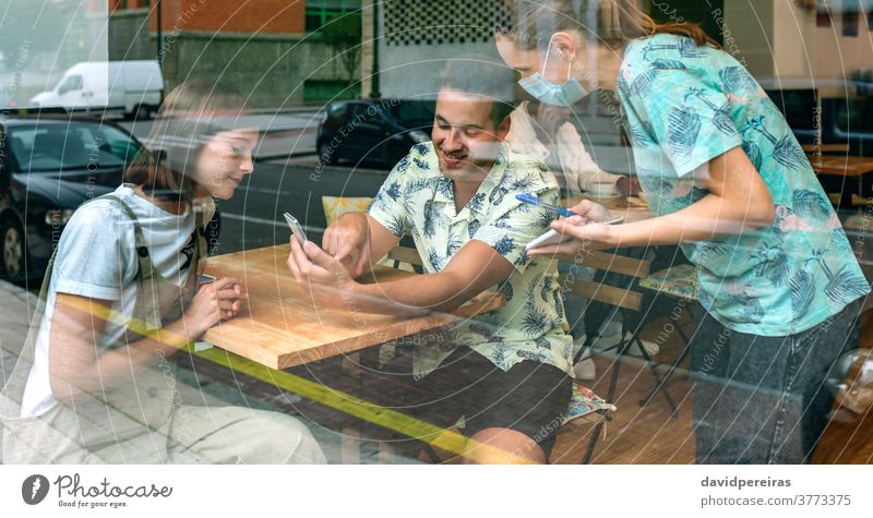 Waitress with mask taking note to couple restaurant menu mobile ordering food covid-19 waitress protective mask smiling asking reflection through glass diner