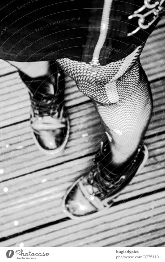Woman photographed from the hip downwards in standing position. She wears a black skirt with a conspicuous zipper and chains, fishnet tights with runners and holes and rough black shoes. The focus is on the big running stitch.