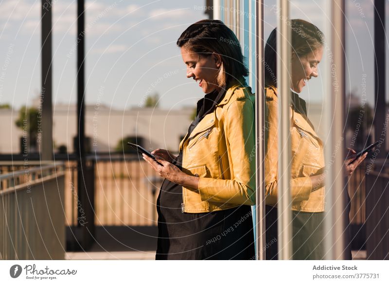 Smiling pregnant woman reading messages on smartphone chat social media city glass wall building sunlight using female tummy belly cheerful smile watch online