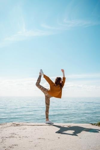 Young woman in a sweater on a beach practicing yoga Dancer's Pose (Natarajasana) yogini street cool training sportswear movement workout outdoors stylish