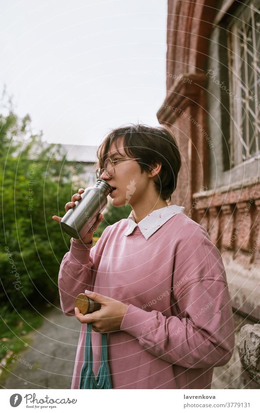 Young adult female with short hair drinking from metal sustainable water outdoors, selective focus bottle hands closeup fingers holding woman girl pink