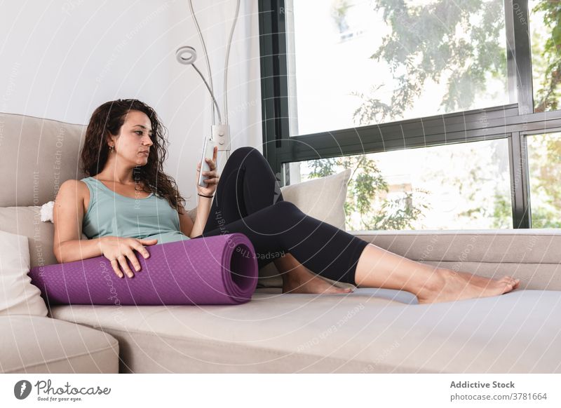 Tranquil woman relaxing on sofa with smartphone and yoga mat browsing home lying using weekend female sportswear activewear living room couch soft modern