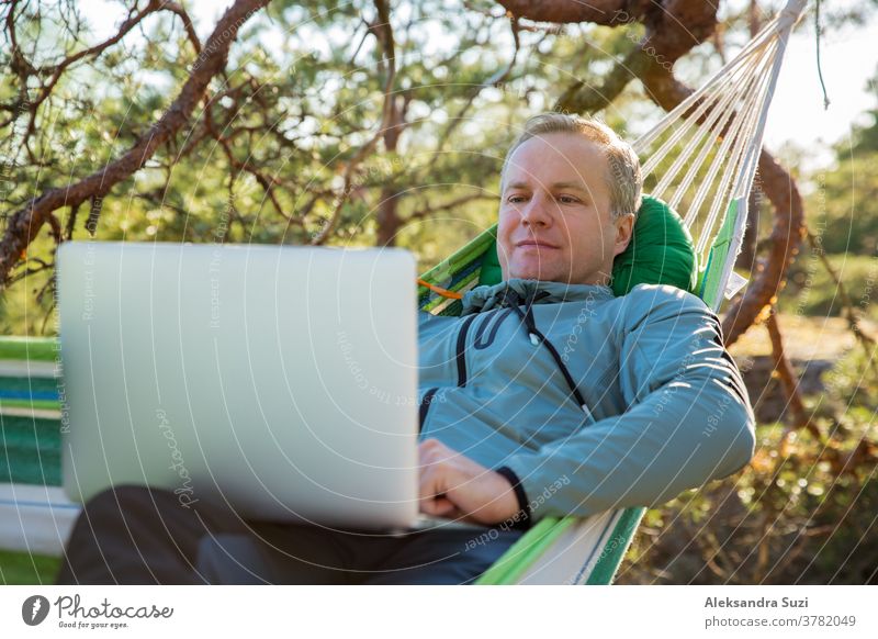 A man working on a laptop while lying in a hammock in the woods. Self-isolation, freelancing, remote work and distancing. Top view of Scandinavian landscape