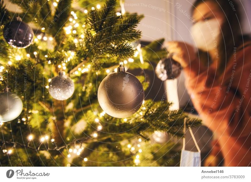 Young woman decorating a Christmas tree with medical safety mask for Covid-19, Coronavirus and Christmas concept, beautiful holiday new year home prevention