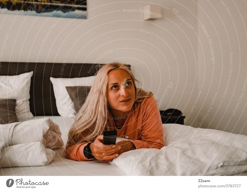 Young woman lies on the bed and zaps through the TV programs Television Remote control Blonde long hairs Bed relaxation Relaxation fortunate cheerful kind