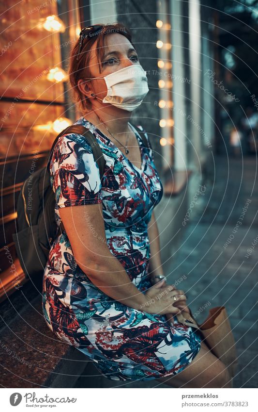Young woman sitting outdoors at coffee shop front after shopping in the city center, wearing the face mask to avoid virus infection caucasian covid-19 lifestyle