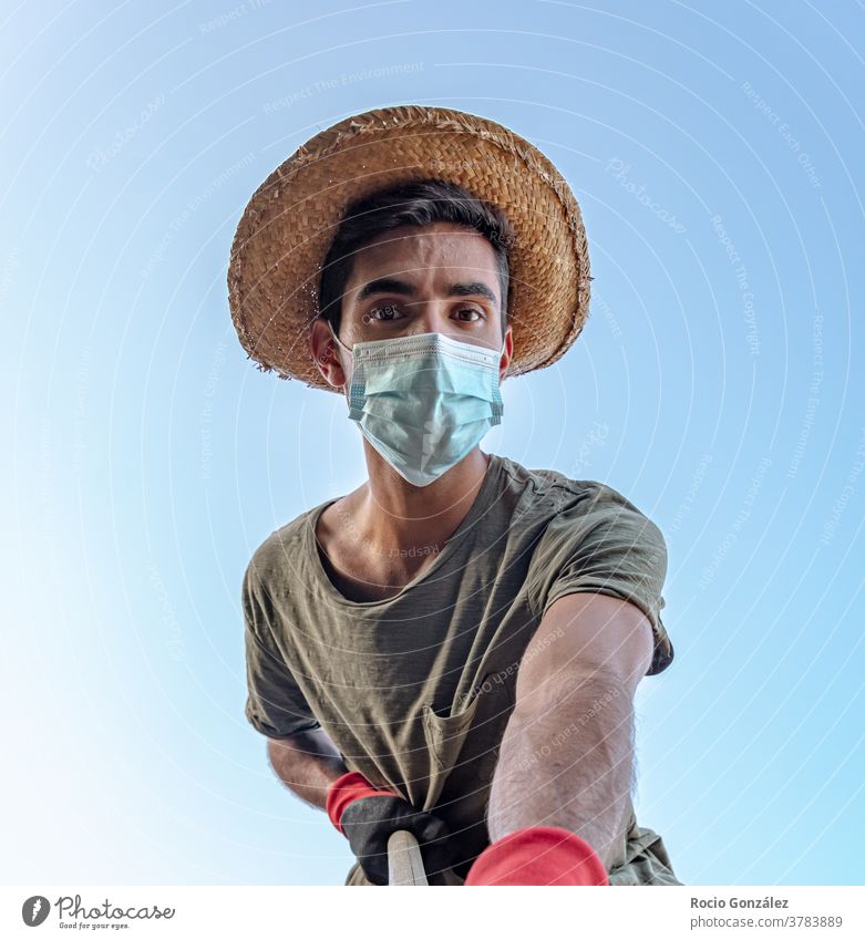 Young man working as a farmer with protection mask in times of covid 19 and seen from below young worker straw hat brunette brunette hair boy gloves Agriculture