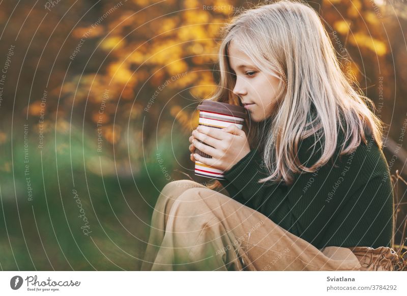 A beautiful teenage girl with blond hair sits thoughtfully in an autumn park, holds a thermos with tea and warms her hands. sad pensive child blonde yellow leaf