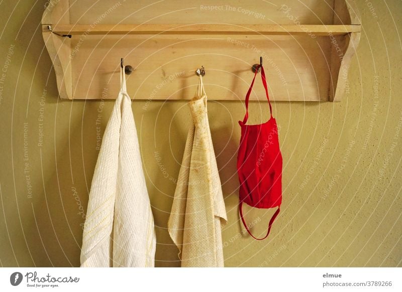 A red nose-mouth mask hangs next to two old, dirty towels on one of the three hooks of a wooden wardrobe Nose and face mask Mouth protection mask Mask Towel