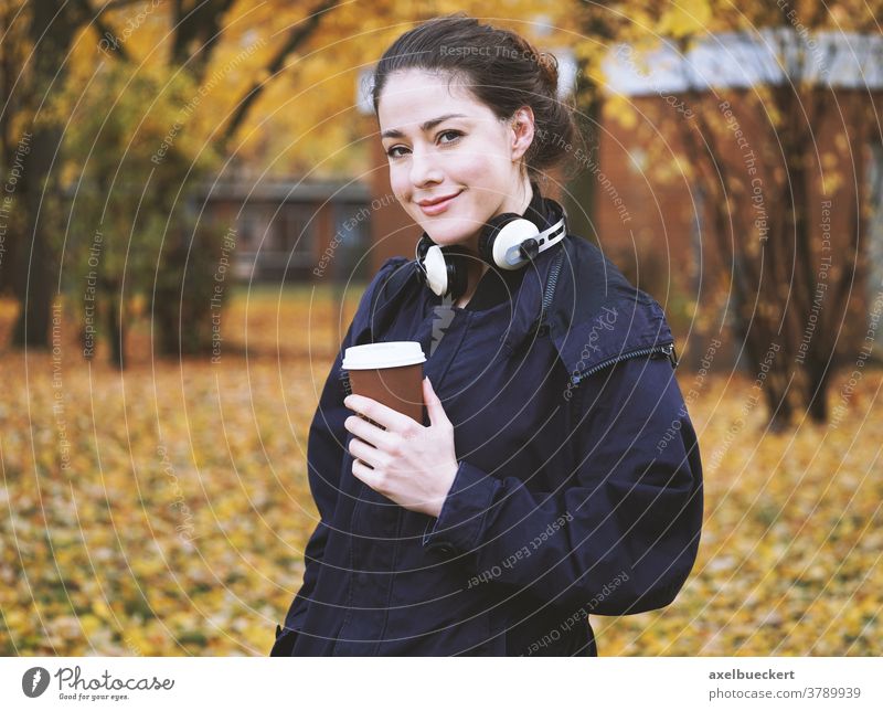 young woman with coffee to go cup and headphones in autumn candid outdoor outside real people fall hot drink cold weather street walk park disposable holding