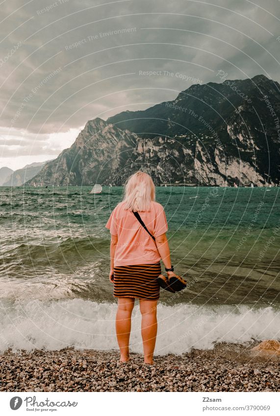Junge Frau steht am Ufer des Gardasees gardasee norditalien torbole urlaub wellen wasser gewässer frisch strand alpen berge wolken himmel blau ferien sommer