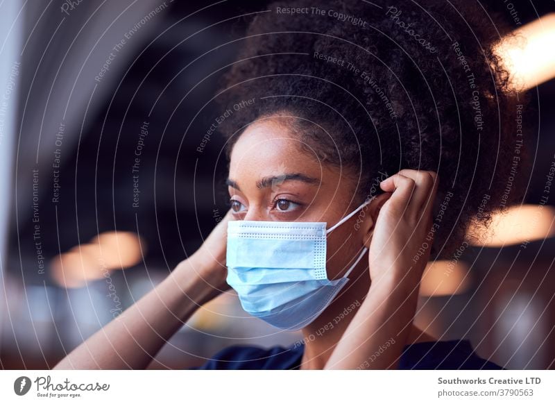 Female Doctor In Scrubs Putting On Face Mask Under Pressure In Busy Hospital During Health Pandemic doctor nurse scrubs key worker female woman putting on