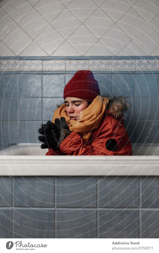 Woman in outerwear sitting in bathtub near tiled wall woman cold eyes closed trendy apparel concept home bathroom glove stylish fur appearance knitted hat scarf