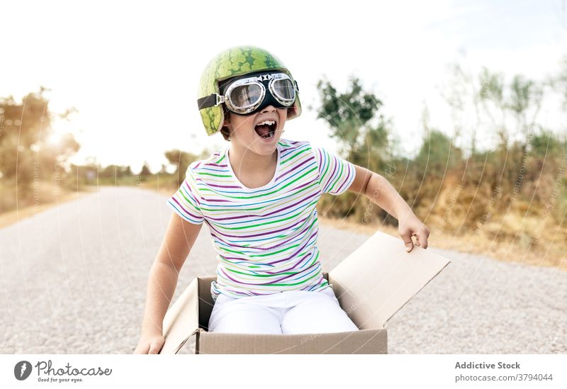Excited boy in goggles sitting in cardboard box on road excited having fun childhood mouth opened stylish carefree sky kid positive smile enjoy stripe wear