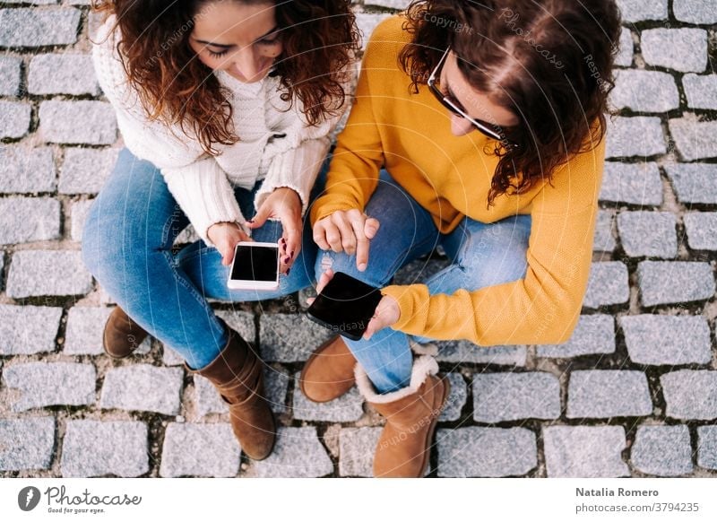 Selective focus. Two beautiful women sitting on the street and using their phones. They are both looking at something on the cell. Technology lifestyle
