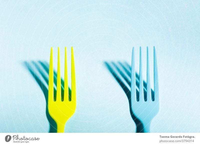Two yellow and blue forks casting a shadow on blue background saturated color isolated surreal concept cutlery silverware food top view still life minimalist