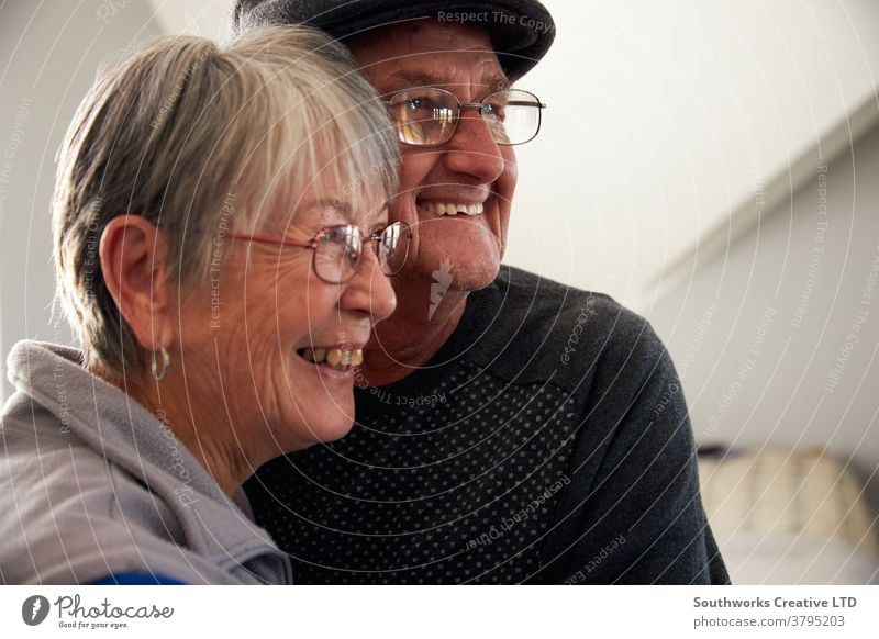 Loving Senior Couple Laughing And Smiling As They Hug Each Other At Home Together couple senior seniors retired at home relaxing standing window loving