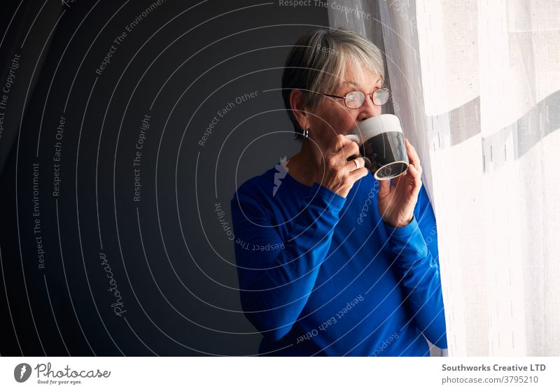 Senior Woman Relaxing Standing By Window At Home With Hot Drink woman senior seniors retired at home relaxing standing window drink drinking hot drink