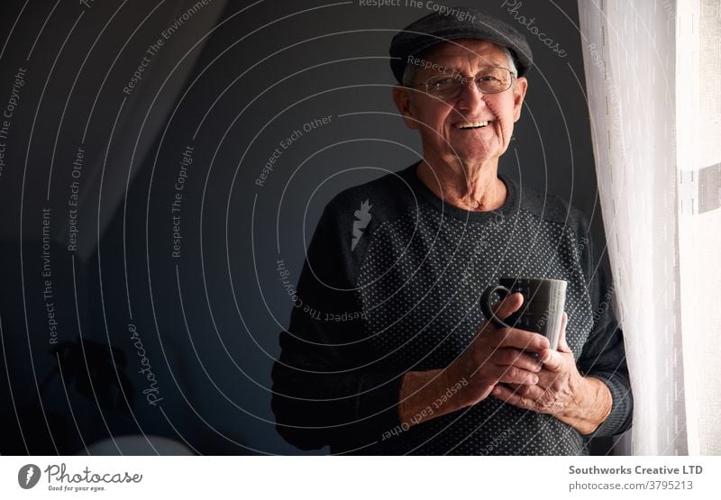 Portrait Of Senior Man Wearing Cap Relaxing Standing By Window At Home With Hot Drink man senior seniors retired at home relaxing standing window drink drinking