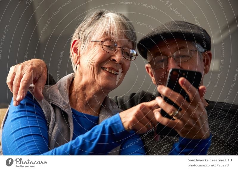 Senior Couple At Home Sitting On Sofa And Using Mobile Phone Together couple senior seniors at home mobile mobile phone cell cell phone connected online tech