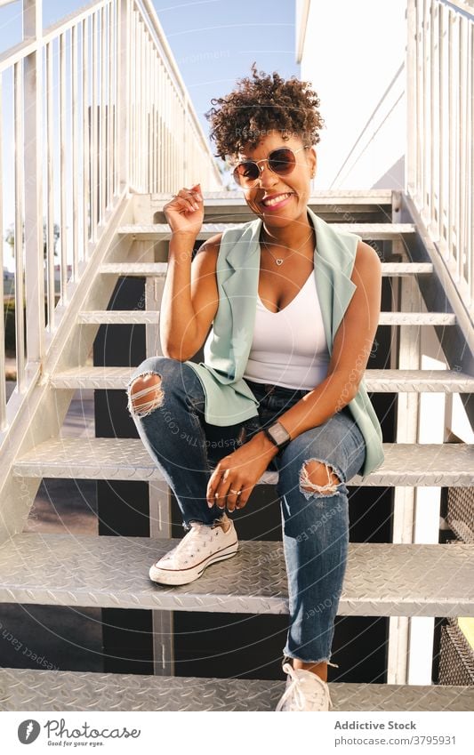 Smiling young black woman sitting on stairway hipster style trendy cheerful happy smile urban sunglasses modern african american ethnic afro outfit female