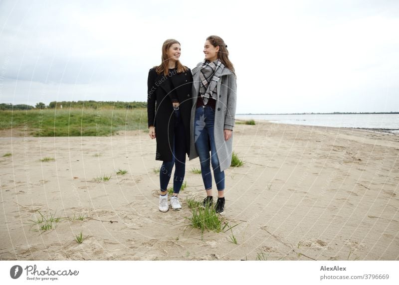 Portrait of two young women - friends - on the beach of the Elbe Woman Young woman 18-25 years warmly pretty Charming Slim Brunette long hairs Fresh Large smart