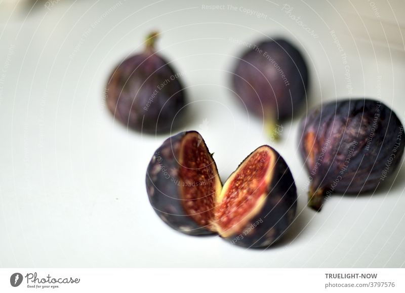 Fresh, ripe figs in a dark blue violet skin, one of them cut open showing its red flesh, lying on a white table in bright daylight Figs Mature fruits Blue Red