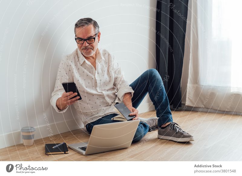 Positive mature man using smartphone and laptop on floor workspace browsing focus positive gadget device watching app concentrate casual at home busy male