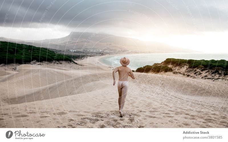 Anonymous sporty naked man running on sandy desolate seacoast nude active carefree freedom beach scenic travel seashore hat fit bare unclothed enjoy tranquil