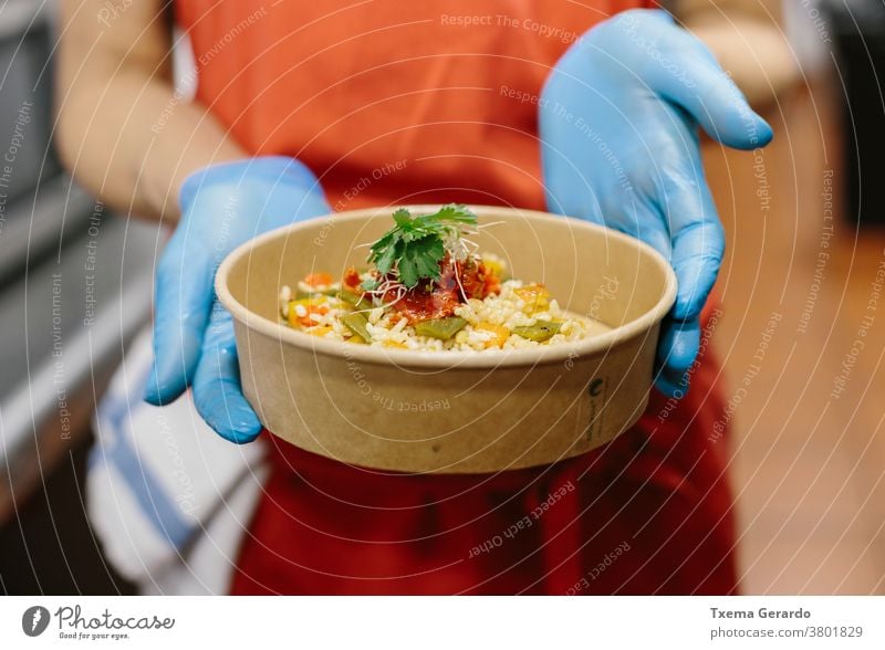 A cook presents her gourmet rice salad in a compostable take-away container takeaway food vegetarian vegan tomatoes beans apron hands gloves kitchen restaurant