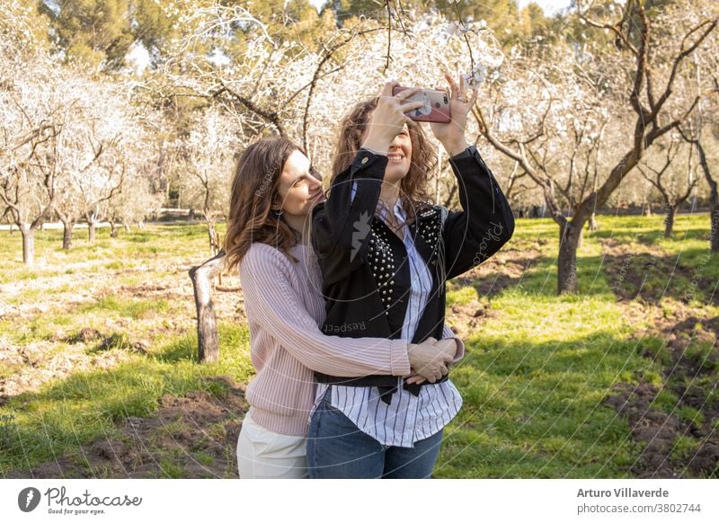 two girls in a park take cell phone selfies while hugging. They are both very pretty and smile happily. One is wearing a pink sweater and the other is wearing a studded jacket