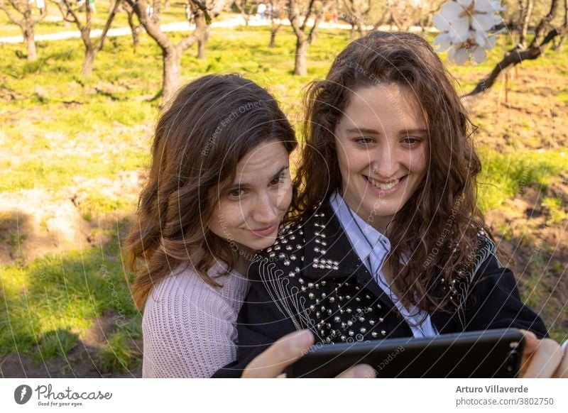 two girls in a park take mobile selfies. They are both very pretty and smile happily. One is wearing a pink sweater and the other is wearing a studded jacket