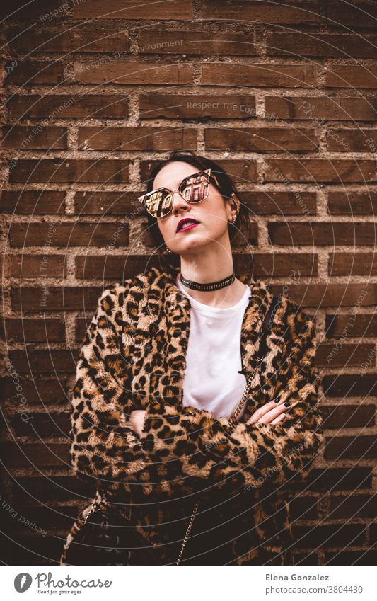 Stylish portrait of young serious model with crossed arms wearing trendy leopartd print faux fur coat, fashion sunglasses, posing on red brick wall texture grunge background