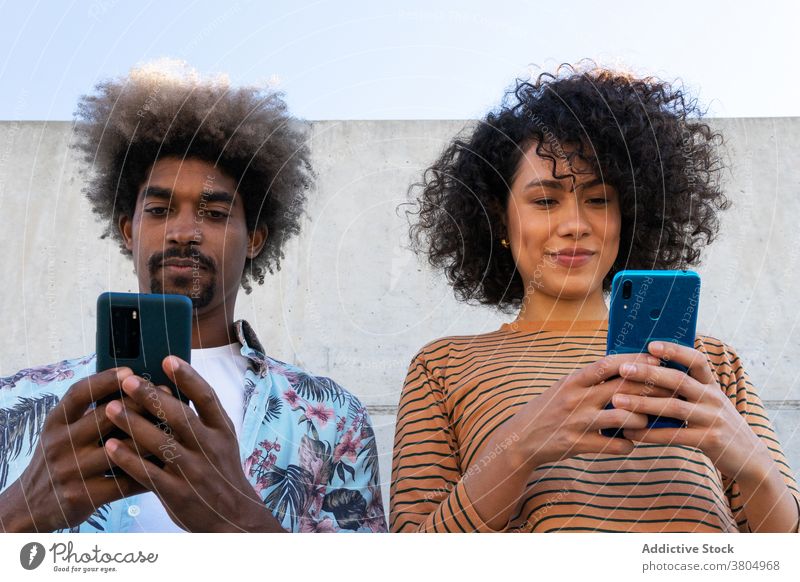 Glad multiethnic friends watching smartphones on street couple content stylish wear hairstyle friendship using gadget cheerful cellphone trendy afro device