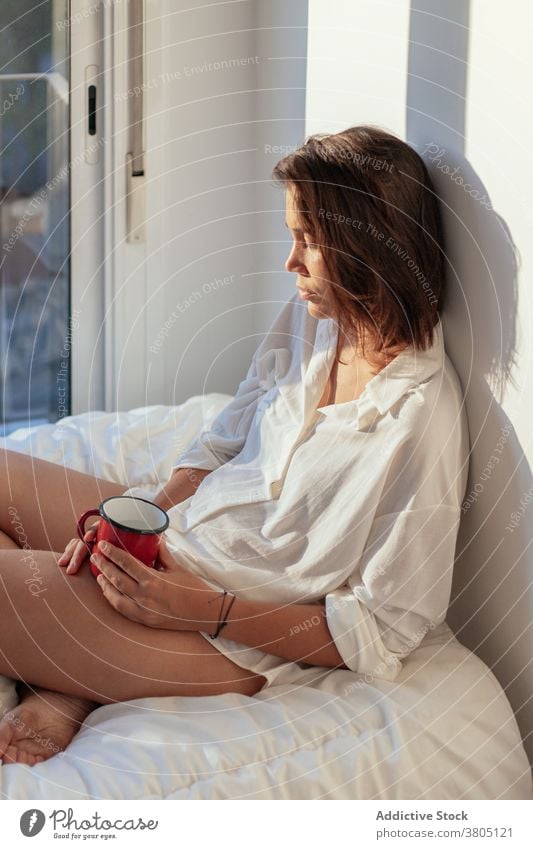 Young woman drinking coffee in bed morning pensive thoughtful sensual comfort allure cozy joy female young white shirt body barefoot home beverage sunny relax
