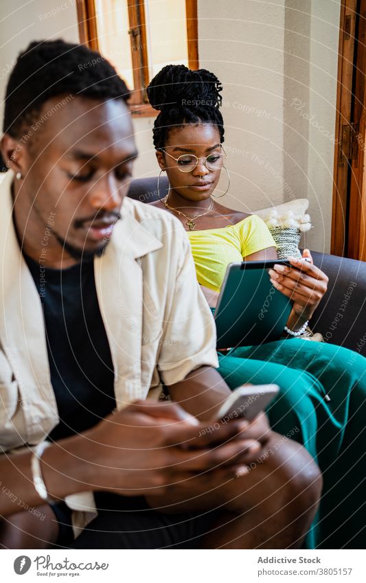 Young ethnic couple using gadgets while relaxing on couch at home sofa together relationship tablet smartphone addict connection browsing online internet