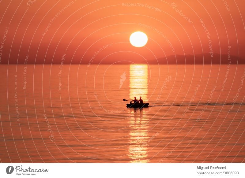 Silhouette of two people in a canoe passing by the reflection of the sun in the sea at sunset. beach silhouette orange kayak horizon browsing boat red landscape