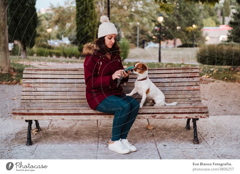 woman sitting on a bench, using mobile phone in a park with her adorable jack russell dog. Lifestyle outdoors autumn brick wall casual clothing caucasian