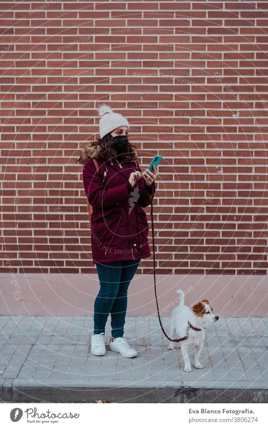 woman wearing protective mask, using mobile phone, walking at the city with her adorable jack russell dog. Lifestyle outdoors allergic autumn brick wall care