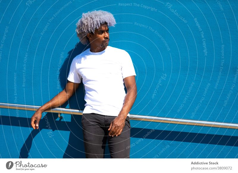 Black man with afro hair dressed in white leaning against a blue black man afro black man afro man leaning on a wall white shirt blue background blue wall