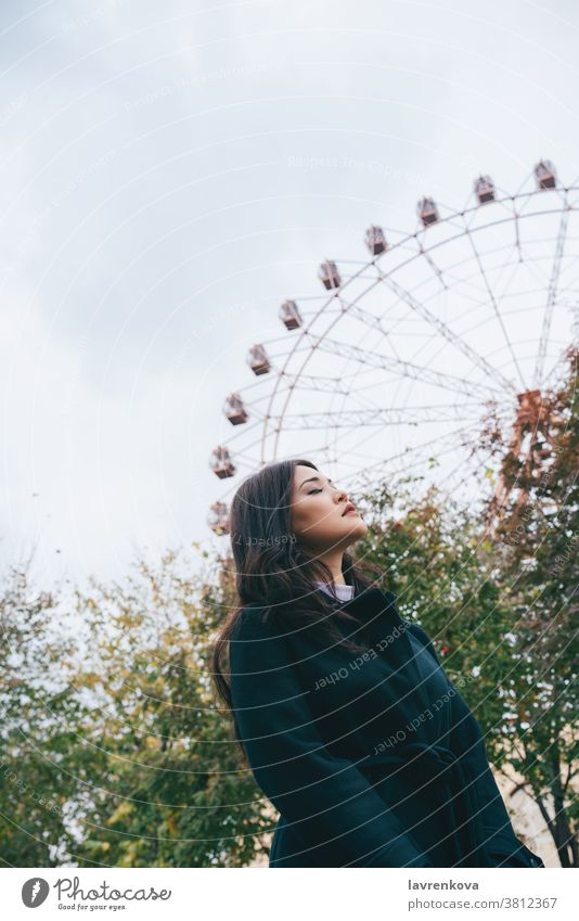 Young adult asian female in a dark coat in front of ferris wheel outdoors, selective focus woman autumn park casual leisure seasonal weekend young walkin fun