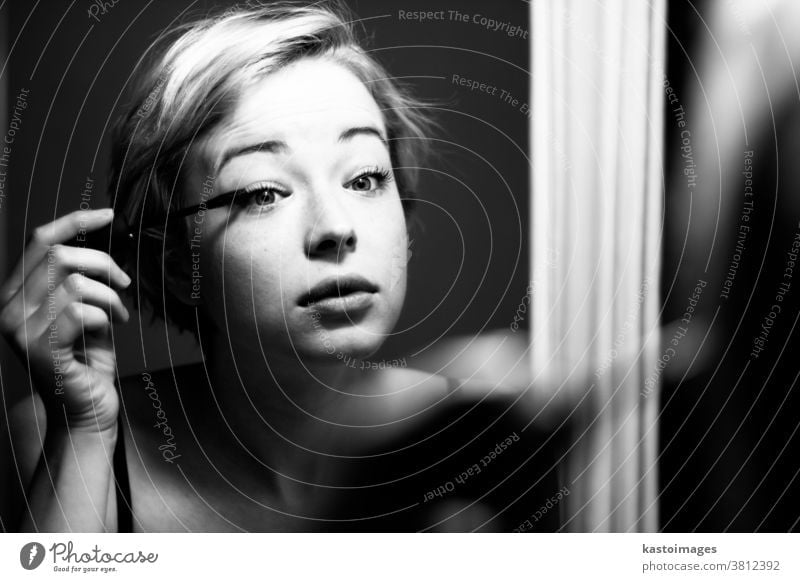 Woman getting ready for work doing morning makeup routine applying mascara in bathroom mirror at home. Beautiful caucasian girl applying eye make-up. Black and white image.