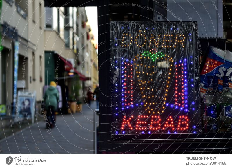 Fast-in-bite gourmet kiosk on the sparsely populated footpath of a shopping street with a brightly lit advertising display that signals the words doner and kebab and a grill with meat skewer