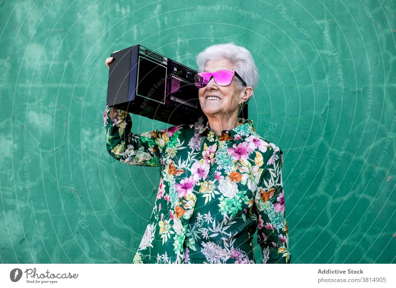Cheerful senior woman in hipster outfit carrying record player trendy music style funny cool elderly female colorful sunglasses fashion listen urban lifestyle