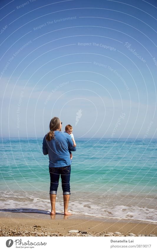 Father and son at the beach baby barefoot calm child childhood copy space day daytime father father and son from behind holding little boy look at the sea man