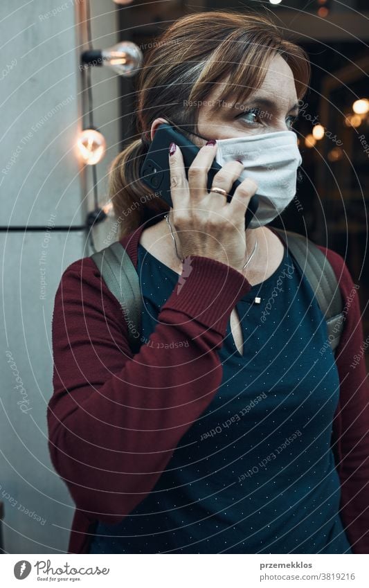 Young woman talking on phone standing in a front of restaurant downtown wearing the face mask to avoid virus infection call care caucasian chat contagious