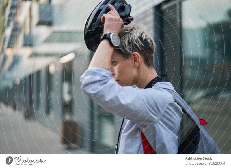 Young woman cyclist puts on helmet while standing next to modern building on summer day in city Woman close-up side view bike walk girl young riding beautiful