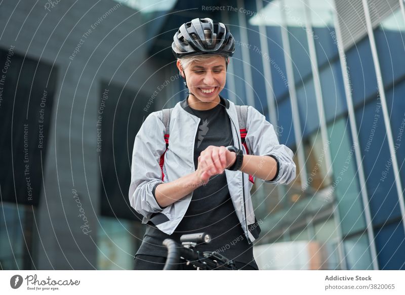 Young woman cyclist looks at wristwatch on background of modern building Cyclist city day young sports riding summer helmet protective happy girl delivery